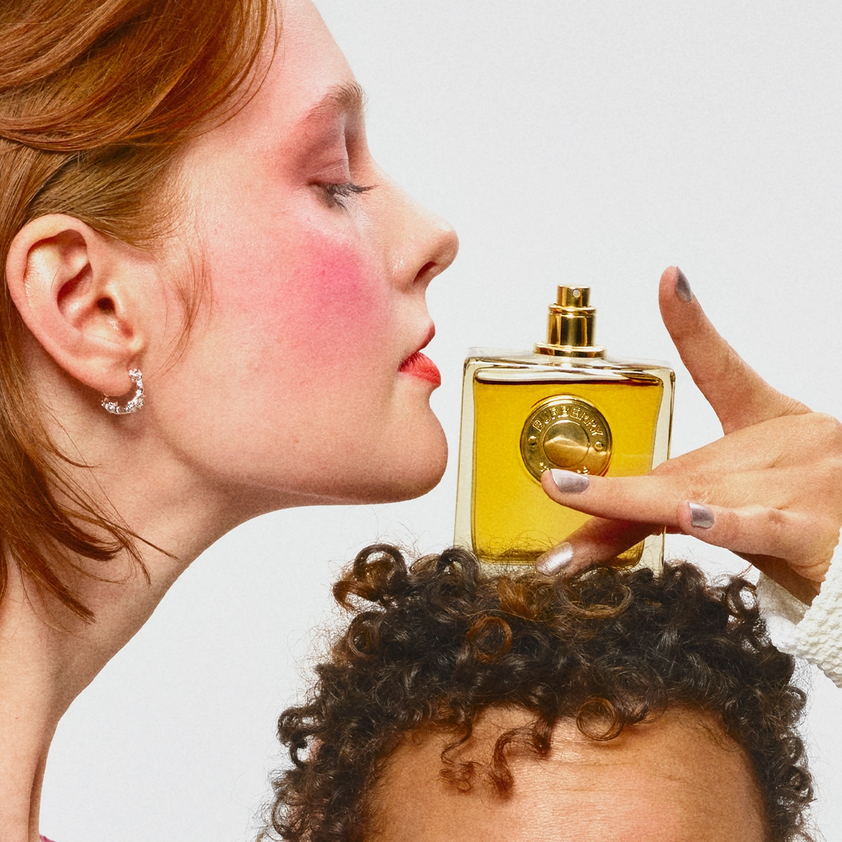 Henkilö kumartuu tuoksumaan keltaista hajuvesipulloa, jota pidetään toisen henkilön kiharan hiuksen yläpuolella. Lähikuvassa hajuvesi keltaisella nesteellä ja pyöreä kultainen etiketti sekä näkyvä poskipuna ja punainen huulipuna.