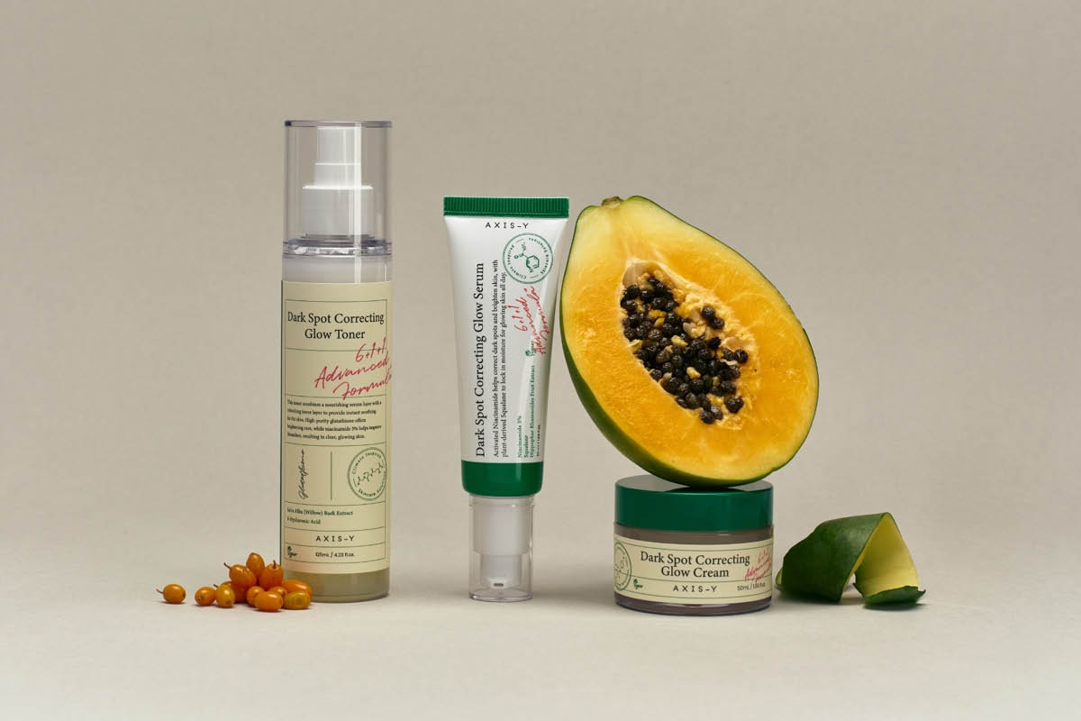 AXIS-Y-ihonhoitotuotteita teksteillä “Dark Spot Correcting Glow Toner”, “Dark Spot Correcting Glow Serum” ja “Dark Spot Correcting Glow Cream”, aseteltuna papaijan ja pienten oranssien marjojen viereen neutraalilla taustalla.