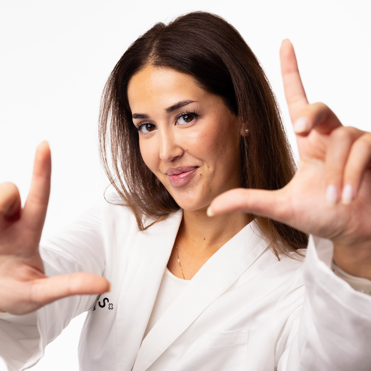 En person i vit arbetsrock håller upp händerna framför kameran och formar en rektangel med fingrarna som en kameraram. Närbild med naturlig makeup, neutral bakgrund och fokus på ansiktet och händerna.