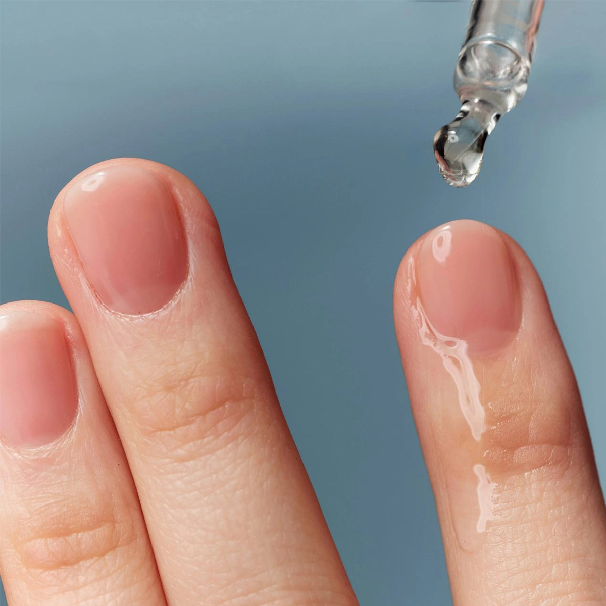 Nærbilde av fingre med gjennomsiktig hudpleieserum som påføres huden med en pipette mot en lyseblå bakgrunn. Fokus på fuktighetsgivende hudpleieserum med glansfull finish og minimalistisk skjønnhetsfølelse.