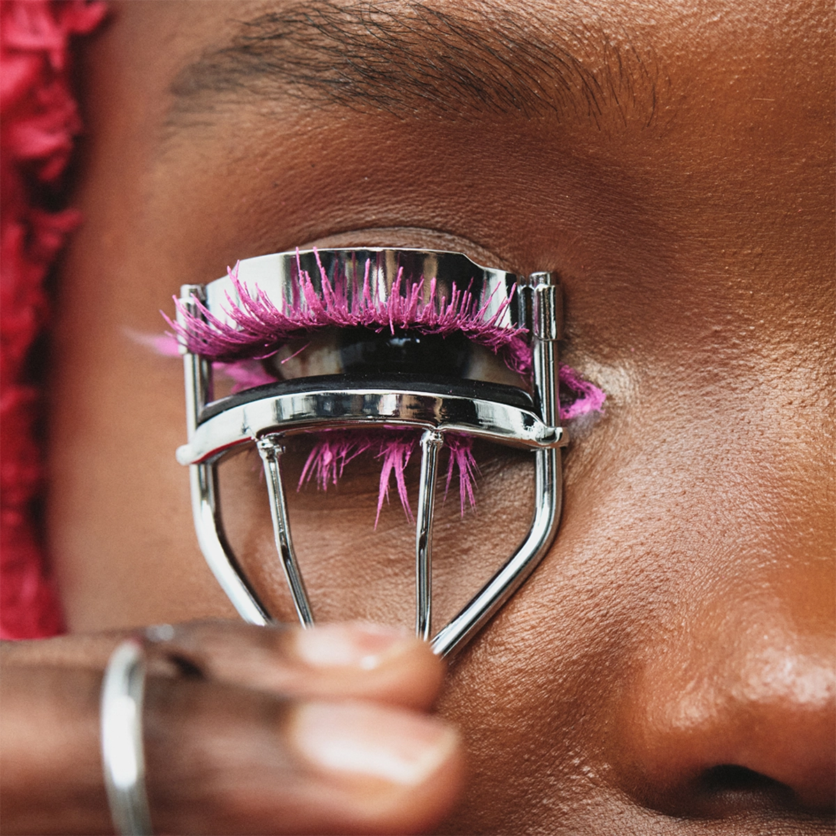 Nærbilde av et øye der en person bruker en øyevippetang på de øvre vippene med rosa mascara, med blanke lepper synlige nederst.