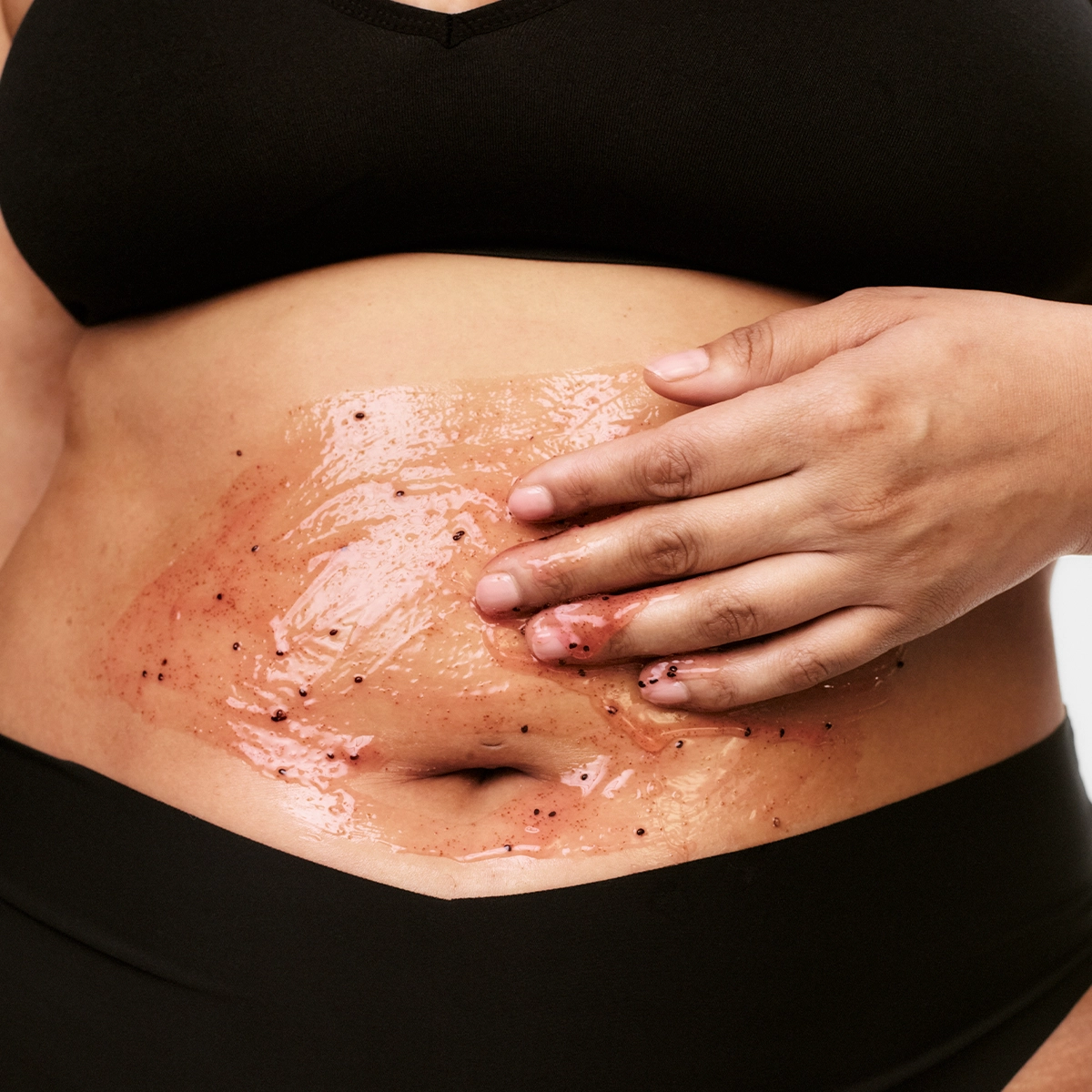 Närbild på en kroppsskrubb med geléliknande peeling och mörka exfolierande korn som appliceras på magen med handen. Hudvårdsprodukt för kroppspeeling och exfoliering med glansig finish på huden.