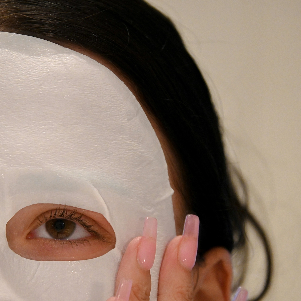 Lähikuva henkilöstä, jolla on valkoinen sheetmask kasvoilla. Henkilöllä on tummat hiukset ja vaaleanpunaiset kynnet. Tausta on beige.
