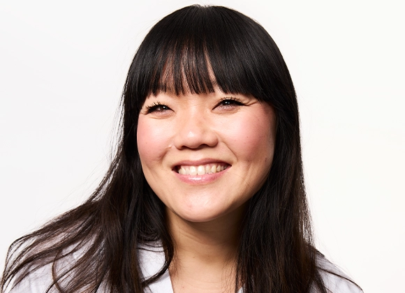 Muotokuva hymyilevästä henkilöstä, jolla on pitkät tummat hiukset ja suora otsatukka, vaaleaa taustaa vasten.