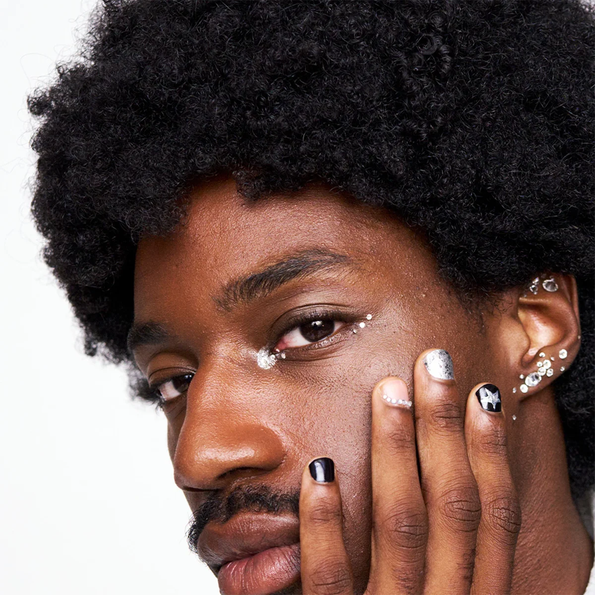 Lähikuvassa näkyy henkilö tummalla ihonsävyllä ja luonnollisilla kiharoilla hiuksilla, kasvot osittain kättä vasten. Kynnet ovat koristeltu mustalla lakalla, hopeisella glitterillä ja taiteellisilla yksityiskohdilla, kuten tähtikuviolla ja pienillä kristalleilla, jotka tuovat ilmeeseen ylellistä ja trendikästä tunnelmaa. Silmien ympärillä näkyy hohtavaa meikkiä sekä poskipäälle aseteltuja pieniä kimaltelevia koristeita, jotka luovat rohkean ja juhlavan lookin. Kuvan kokonaisuus huokuu modernia kauneutta, luovaa itseilmaisua ja näyttävää kynsityyliä.