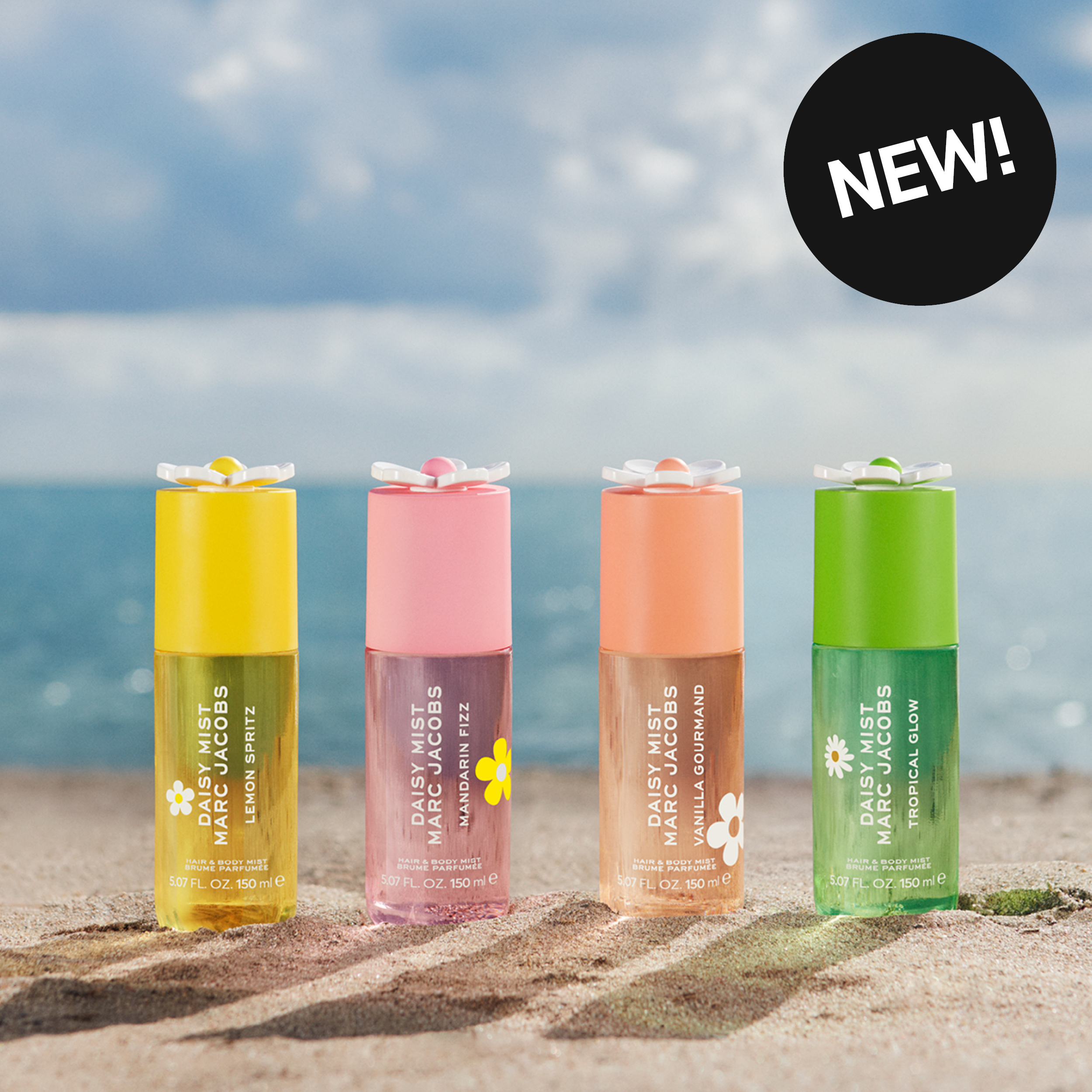 Fyra Daisy Mist by Marc Jacobs hair & body mist står på sand framför havet. Produkterna heter Lemon Spritz, Mandari Fizz, Vanilla Gourmand och Tropical Glow, alla 150 ml, och en svart etikett med texten "NEW!" syns i övre högra hörnet.