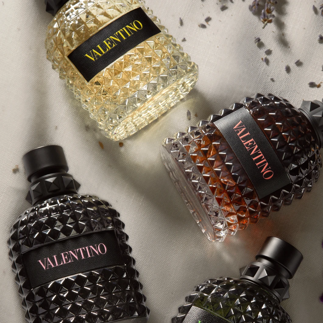 Fire Valentino parfymeflasker med nagledetaljer i ulike farger ligger på et lyst stoff sammen med tørkede blomster. Hver flaske har den synlige teksten "VALENTINO" på etiketten.