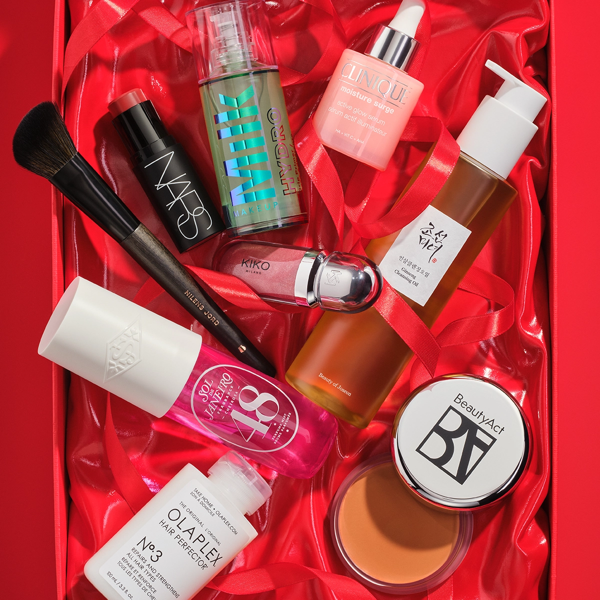 Bild som visar flera skönhetsprodukter arrangerade i en röd presentförpackning. Produkterna inkluderar en svart flaska med NARS foundation, en brun flaska med SKIN VEIL, en rosa flaska med Clinique Moisture Surge, en vit flaska med pump, en burk med BeautyAct bronzer, en flaska med Olaplex Hair Perfector No.3, en makeupborste, och flera andra skönhetsprodukter. Bakgrunden är röd.