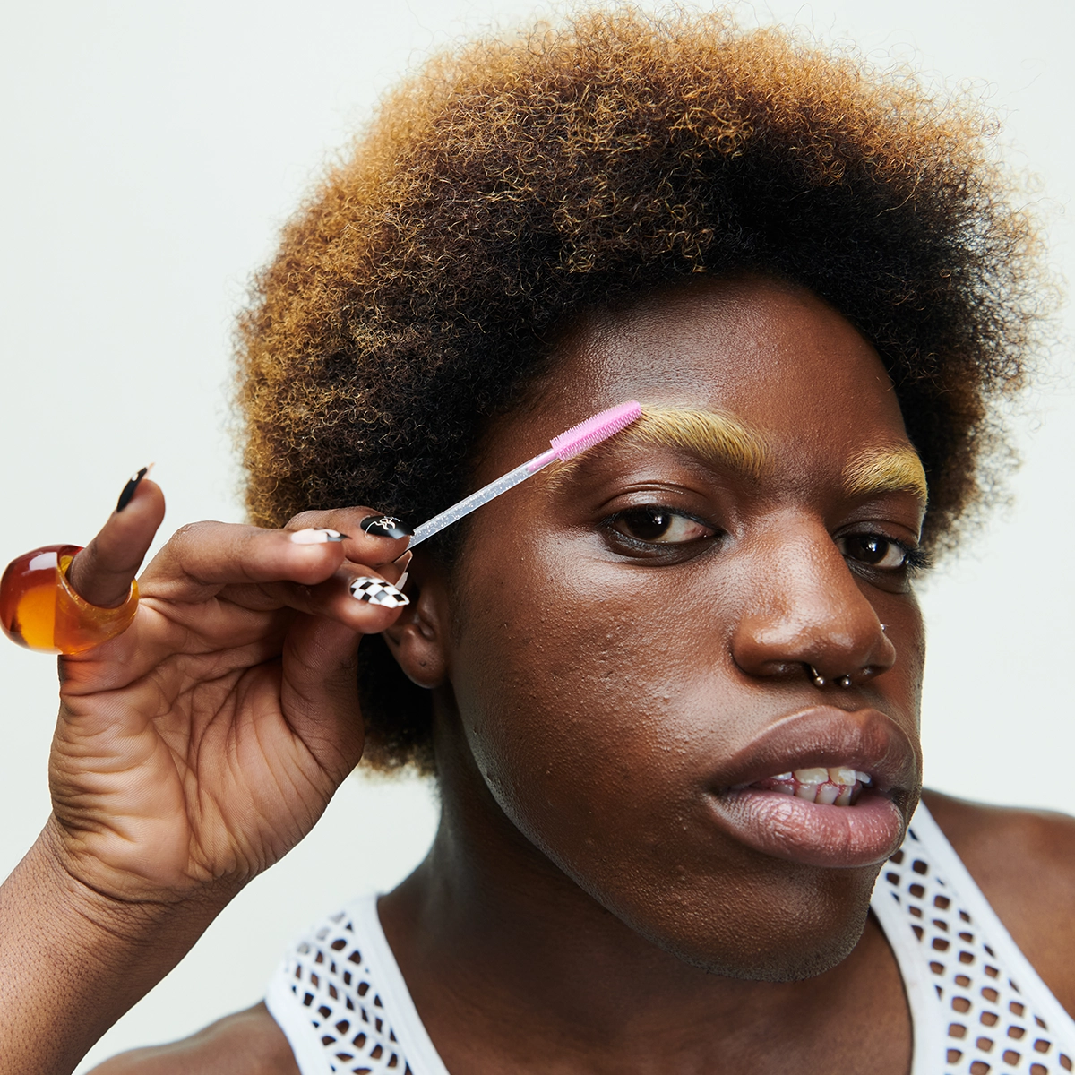 Nærbilde av en person som påfører brynsgel med en rosa spooliebørste på blekede øyenbryn, med fokus på brynsminke og hudtekstur. Naturlig makeup-look med definerte bryn og nøytral bakgrunn.