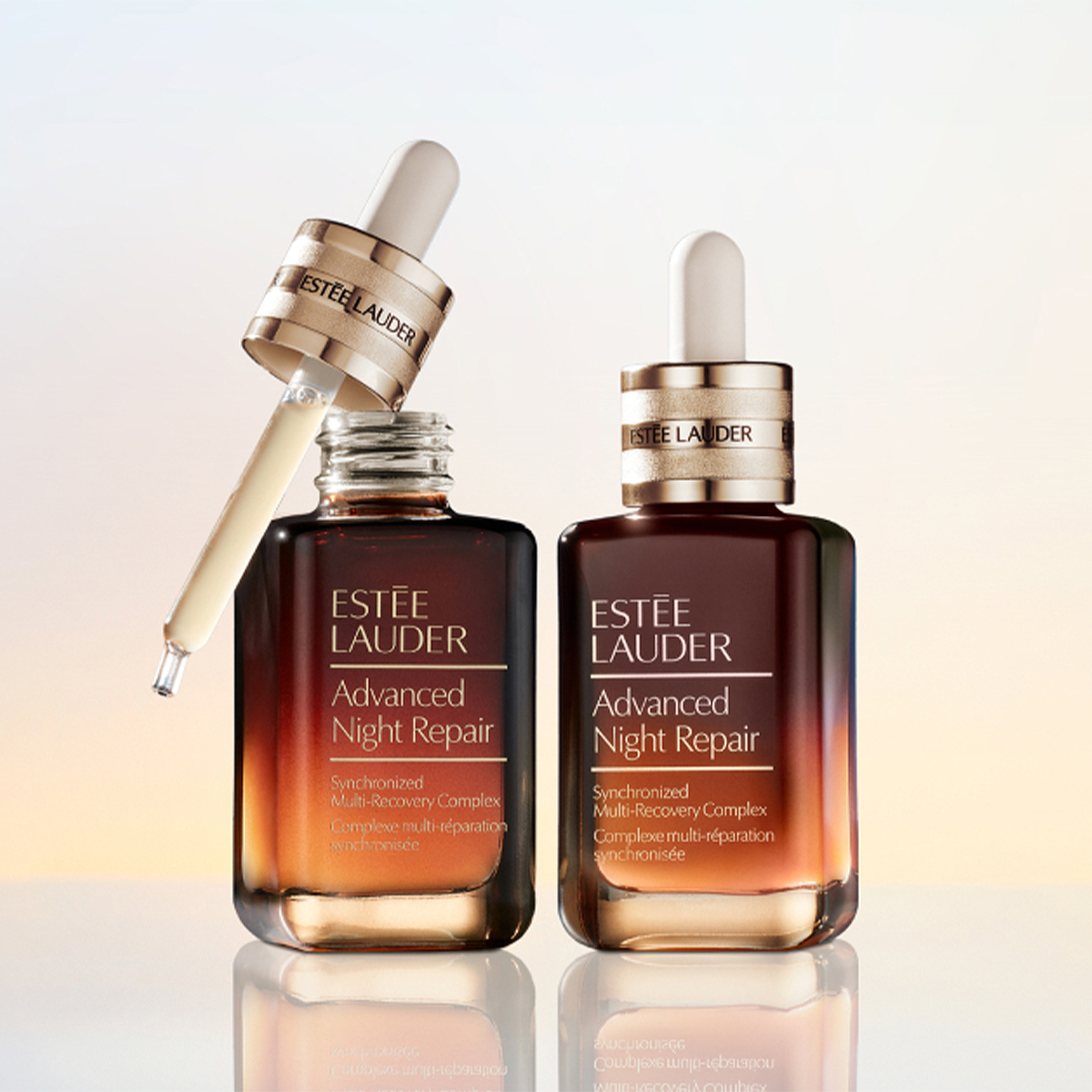 Kaksi Estée Lauder Advanced Night Repair -seerumipulloa on aseteltu heijastavalle pinnalle vaaleaa taustaa vasten. Toinen pulloista on avoinna ja siinä näkyy seerumilla täytetty pipetti sekä teksti ”ESTÉE LAUDER Advanced Night Repair Synchronized Multi-Recovery Complex”, kun taas toisessa suljetussa pullossa näkyy sama tuotteen nimi.
