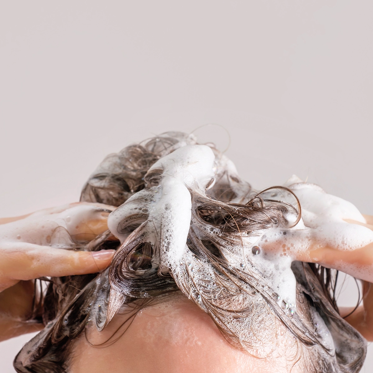 Närbild på en person som tvättar håret med schampo, händer masserar hårbotten och blött hår täckt av vitt lödder mot ljus bakgrund.