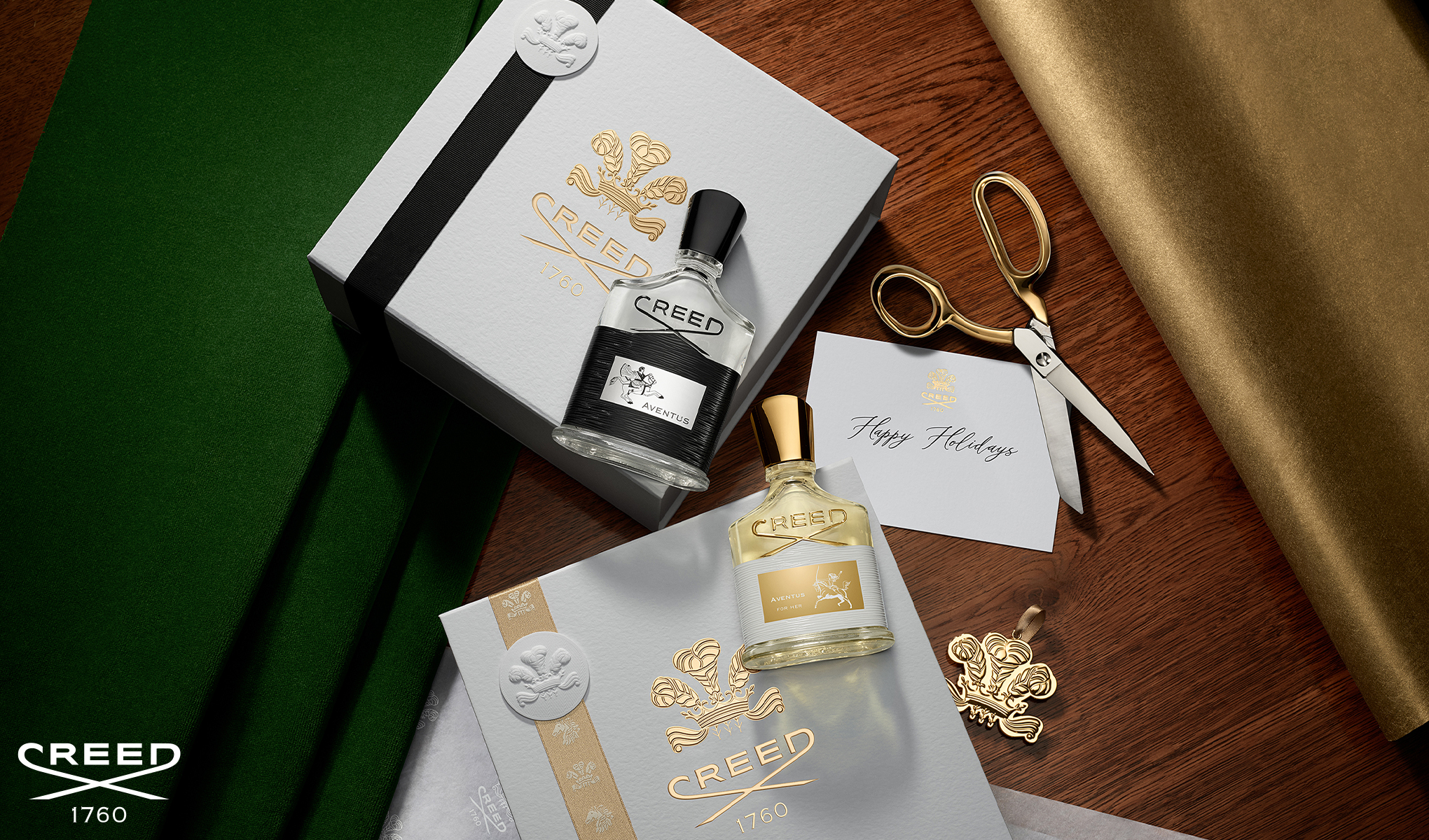 Två Creed Aventus-parfymflaskor ligger i presentförpackningar. I bakgrunden finns grönt och guld presentpapper, en sax och ett gratulationskort.