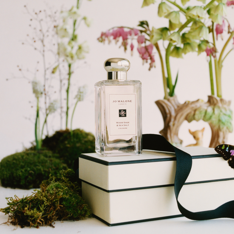 En parfymeflaske fra Jo Malone med teksten «Wood Sage & Sea Salt Cologne» plassert på en hvit gaveeske med et svart bånd. Bakgrunnen er dekorert med grønne planter, mose og blomster.