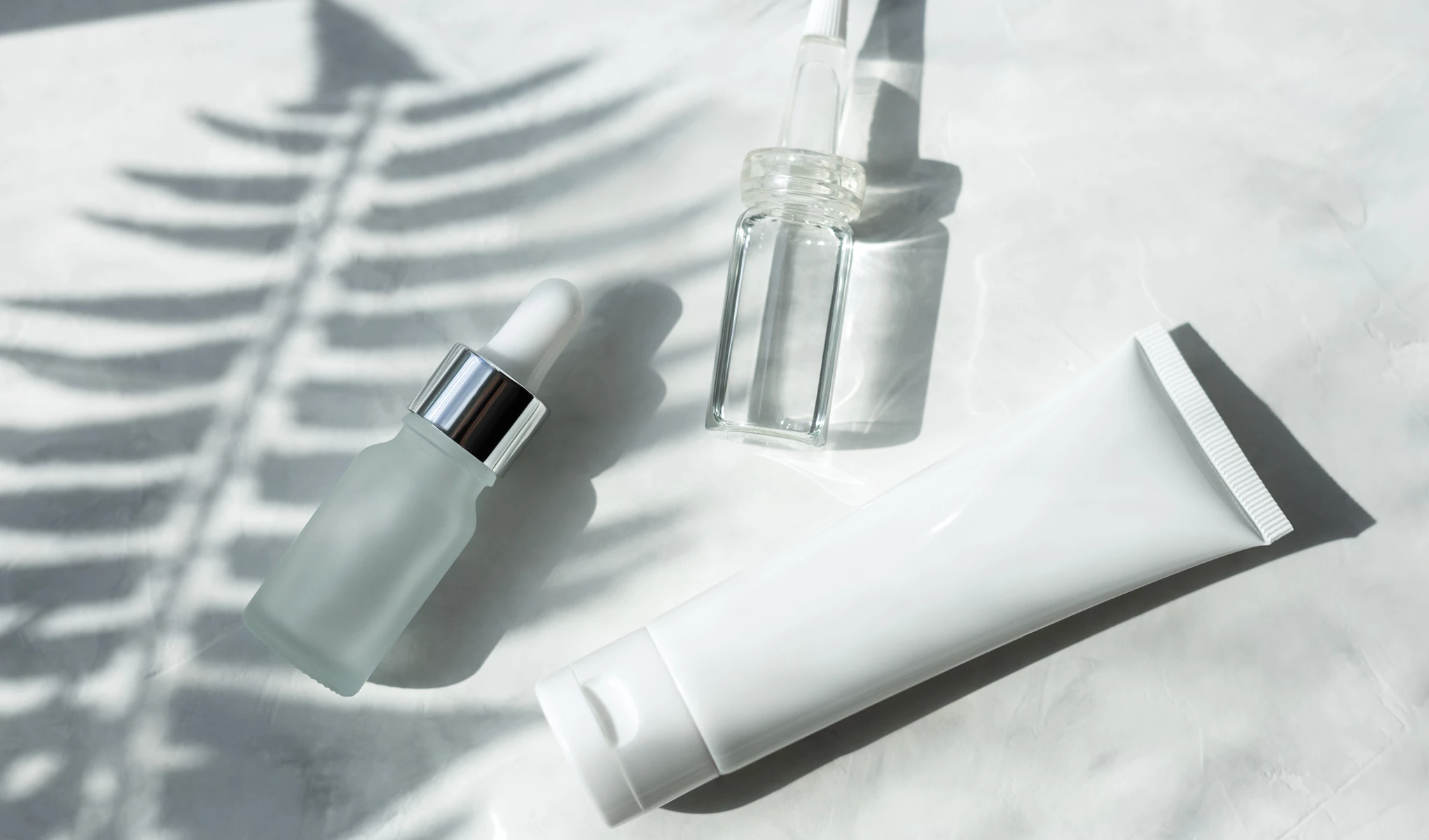 Två obrandade glasflaskor och en vit plasttub ligger mot vit solig bakgrund med skugga av palmlöv.