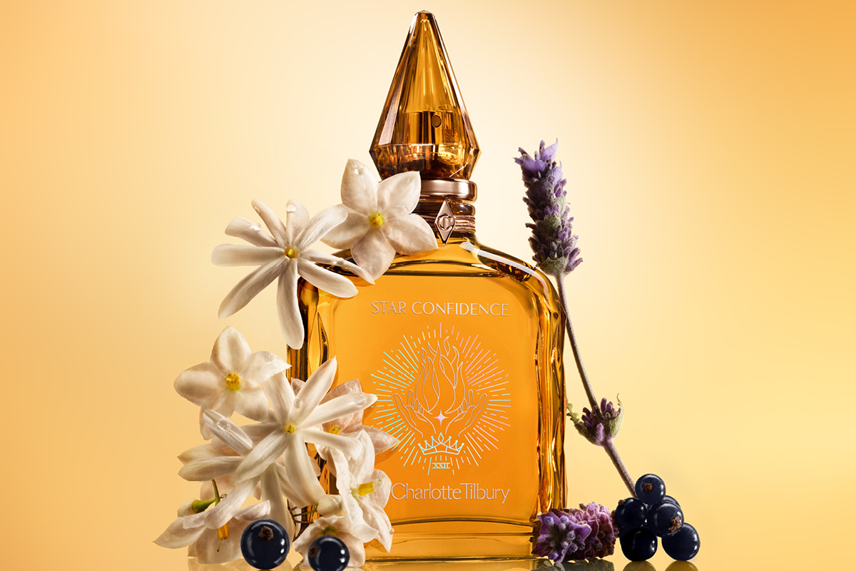 Gyllene parfymflaska (Charlotte Tilbury Star Confidence) omgiven av vita blommor, lavendel och bär mot ljus bakgrund.