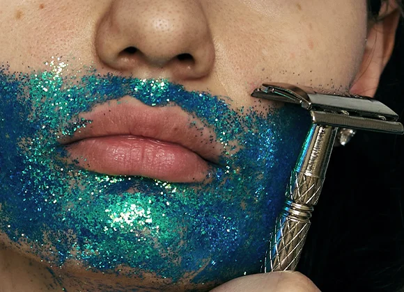 Nærbilde av munn og hake med blågrønt glitter på huden, der en metallbarberhøvel holdes mot haken under barbering mot mørk bakgrunn.