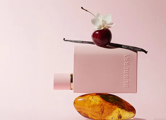 En rektangulär, ljusrosa flaska med texten ”BURBERRY” liggande horisontellt. Flaskan balanserar på en bärnstensfärgad sten. Ovanpå flaskan ligger en mörk vaniljstång, ett rött bär och en vit blomma. Bakgrunden är mjukt rosa och enfärgad.