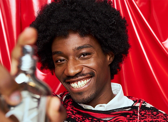 Kuva, joka näyttää henkilön, jolla on tummat kiharat hiukset ja joka pitää hajuvettä. Henkilöllä on valkoinen paita ja punainen ja musta neule. Tausta on punainen.