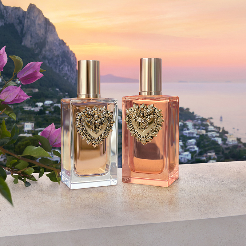 To parfymer fra Dolce & Gabbana, plassert på en steinlignende overflate. Flaskene har et dekorativt hjerteemblem og er fylt med gylden og rosa væske. Bakgrunnen viser en vakker kystby i solnedgang, med havet og fjellene i det fjerne. Blomster med rosa kronblader er synlige til venstre i bildet.