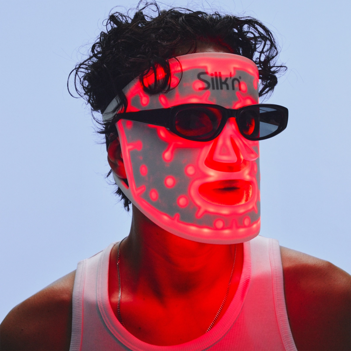 Person bruker en opplyst LED-ansiktsmaske for hudpleie fra Silk’n sammen med svarte solbriller. Masken lyser rødt med synlige lyspunkter og dekker hele ansiktet mot en lys bakgrunn.