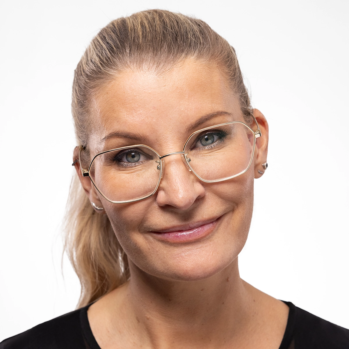 Nærbilde av en person med briller og håret satt opp som smiler svakt mot kamera, med naturlig sminke og jevn hud. Fokus på ansikt, hudpleie og diskret makeup mot nøytral bakgrunn.