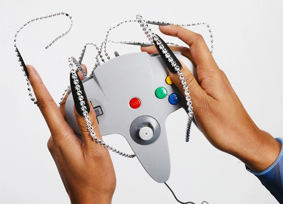 Kaksi kättä pitelee harmaata peliohjainta, jossa on värillisiä painikkeita. Käsien ja ohjaimen päällä on hopeanvärisiä ketjuja ja kimaltavia koristeita vaaleaa taustaa vasten.