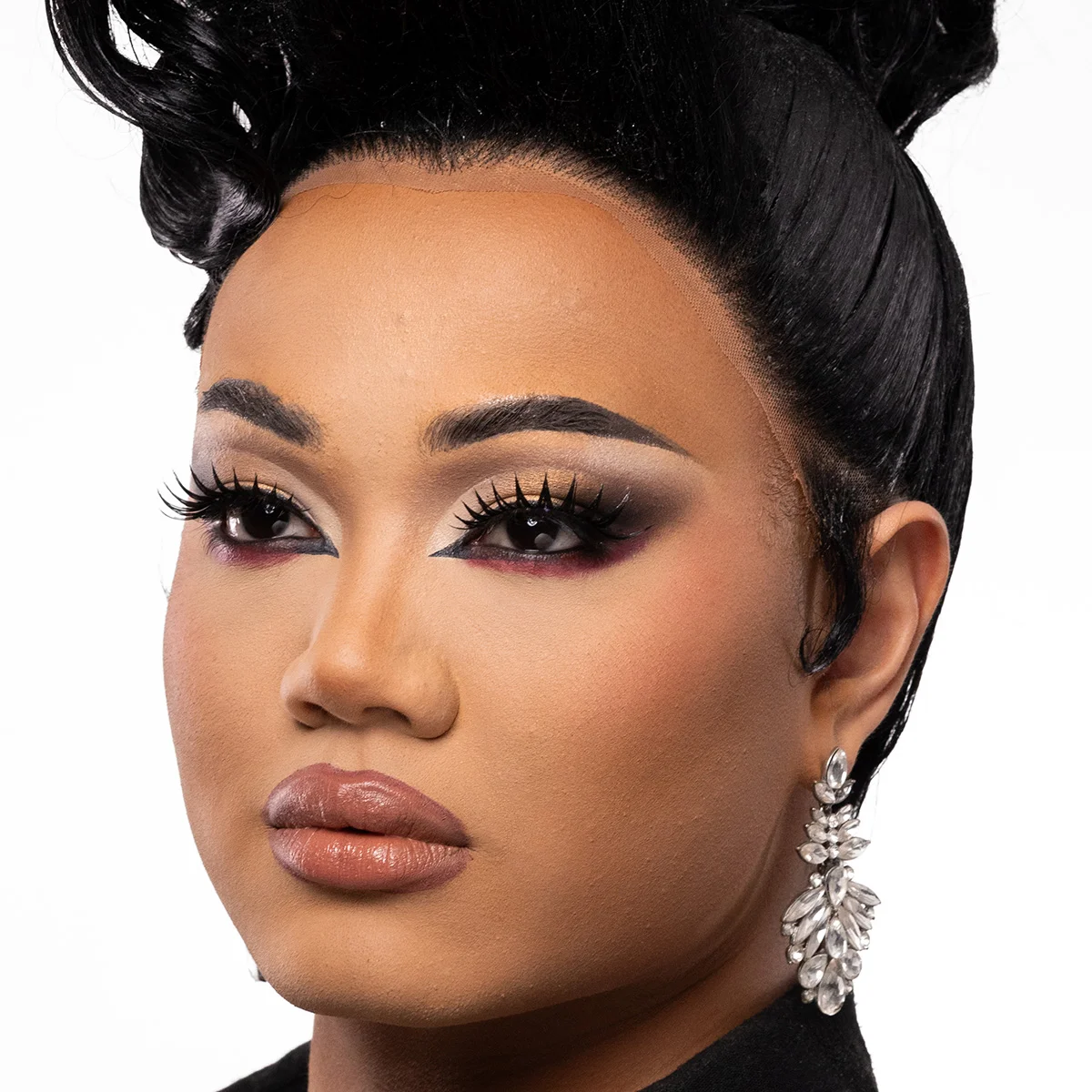 En person med en elegant makeuplook som kombinerar markerad ögonmakeup i guld, svart och vinrött med långa fransar, skulpterad bas och neutrala läppar, kompletterat med stora glittrande örhängen och en uppsatt frisyr.