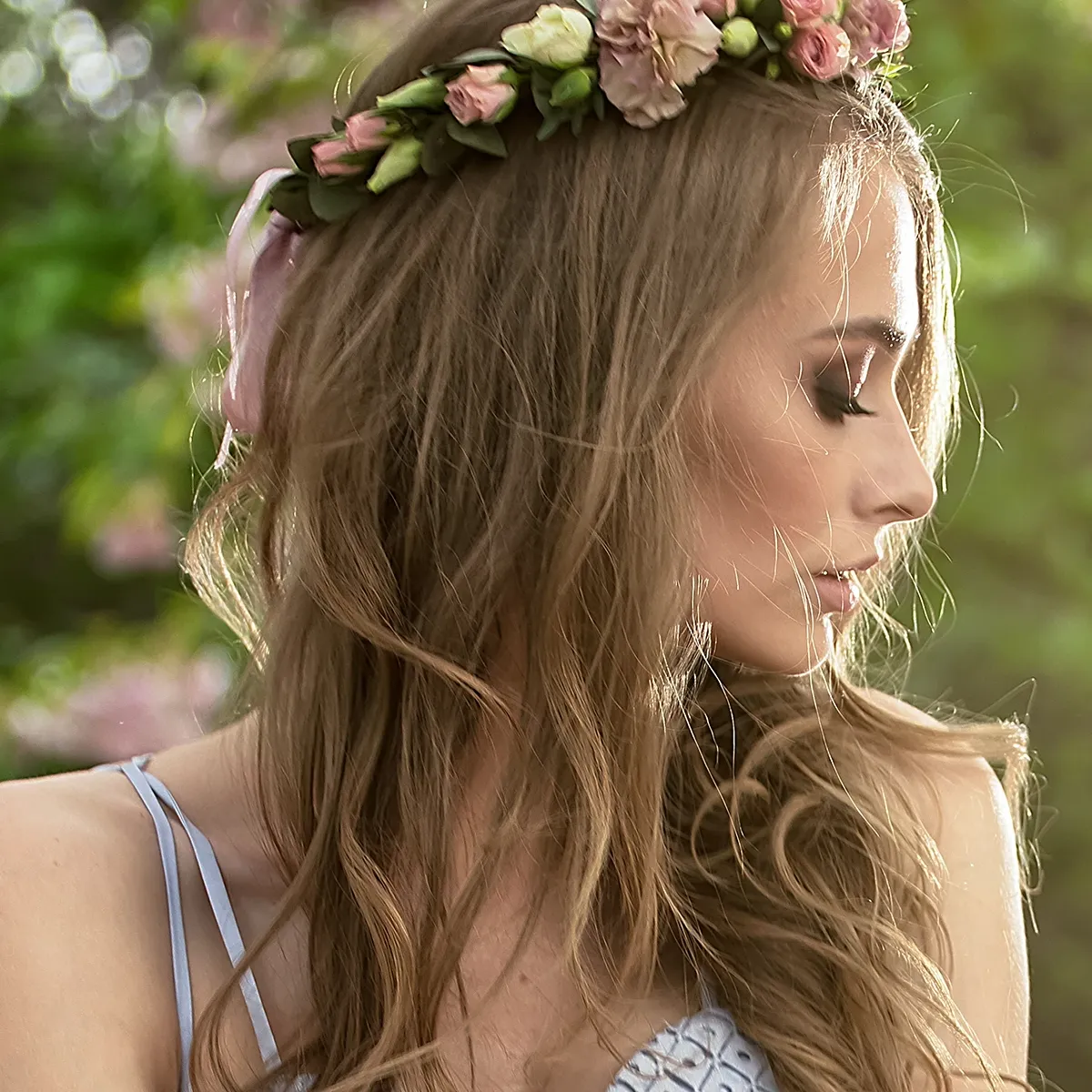 En kvinna med långt hår och en blomkrans på huvudet, i profil framför gröna träd.