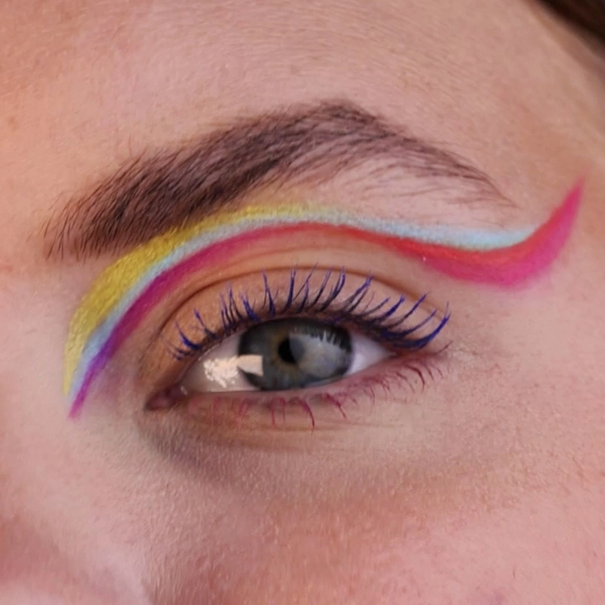 Närbild av ett öga med färgglad eyeliner i flera nyanser längs ögonlocket, inklusive gult, rosa och ljusblått. Ögonfransarna är markerade och bakgrunden är neutral.