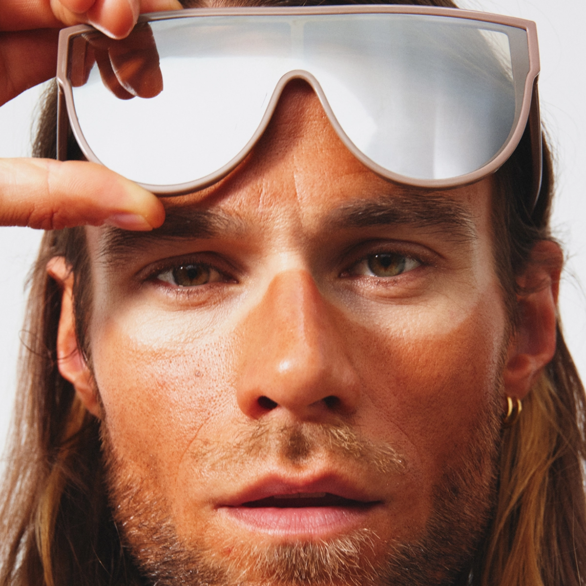 Lähikuva kasvoista, jossa henkilö nostaa aurinkolasit ja osoittaa otsassa näkyvää vaaleaa raitaa aurinkolasien kohdalta. Iholla näkyy epätasainen itseruskettavan sävy ja vaaleampi alue silmien ympärillä.