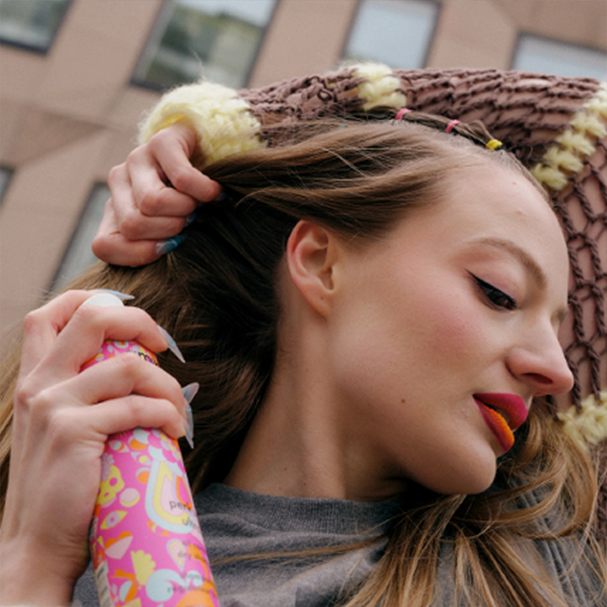 Henkilö suihkuttaa kuivashampoota hiuksiin. Pitkät hiukset, vahva meikki ja värikkäät yksityiskohdat kaupunkiympäristössä.