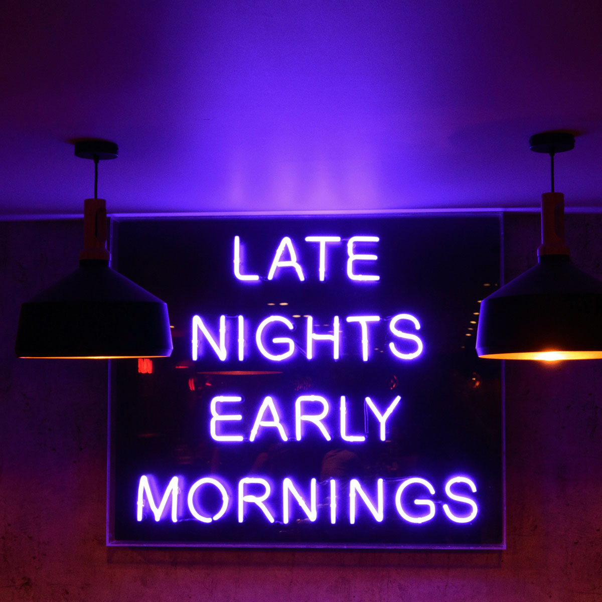 Neonlysskilt med teksten "Late Nights Early Mornings" i et mørkt rom, opplyst av to hengende lamper