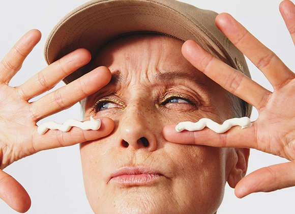 Portrett av en person mot lys bakgrunn som holder begge hendene foran ansiktet. En lys krem er påført i bølgeform under begge øynene. Personen bruker caps og har sminke med gullfarget øyenskygge. Fingrene er strukket ut på hver side av ansiktet.