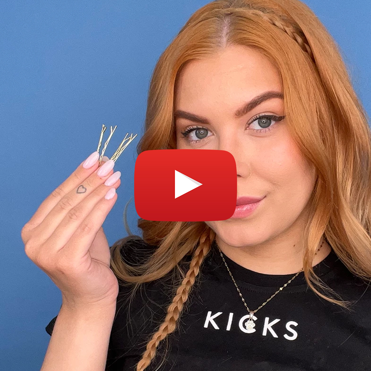 Person med rött hår håller i flera hårnålar. Personen har en fläta i håret och bär en svart tröja med texten 'KICKS'