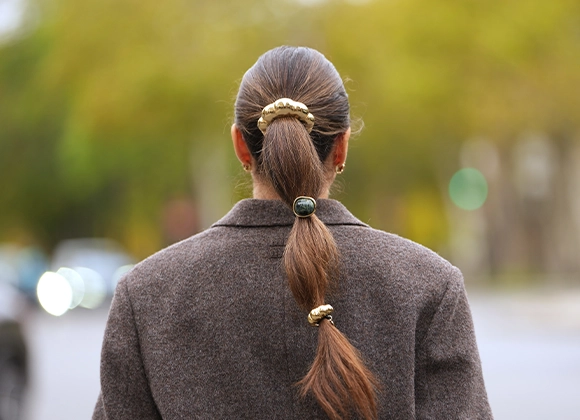 Henkilö nähtynä takaapäin, pitkät hiukset sidottuna matalaksi poninhännäksi useilla hiuslenkeillä. Henkilöllä on tumma takki ja hän seisoo ulkona kadulla, taustalla epätarkkoja puita ja liikennettä.