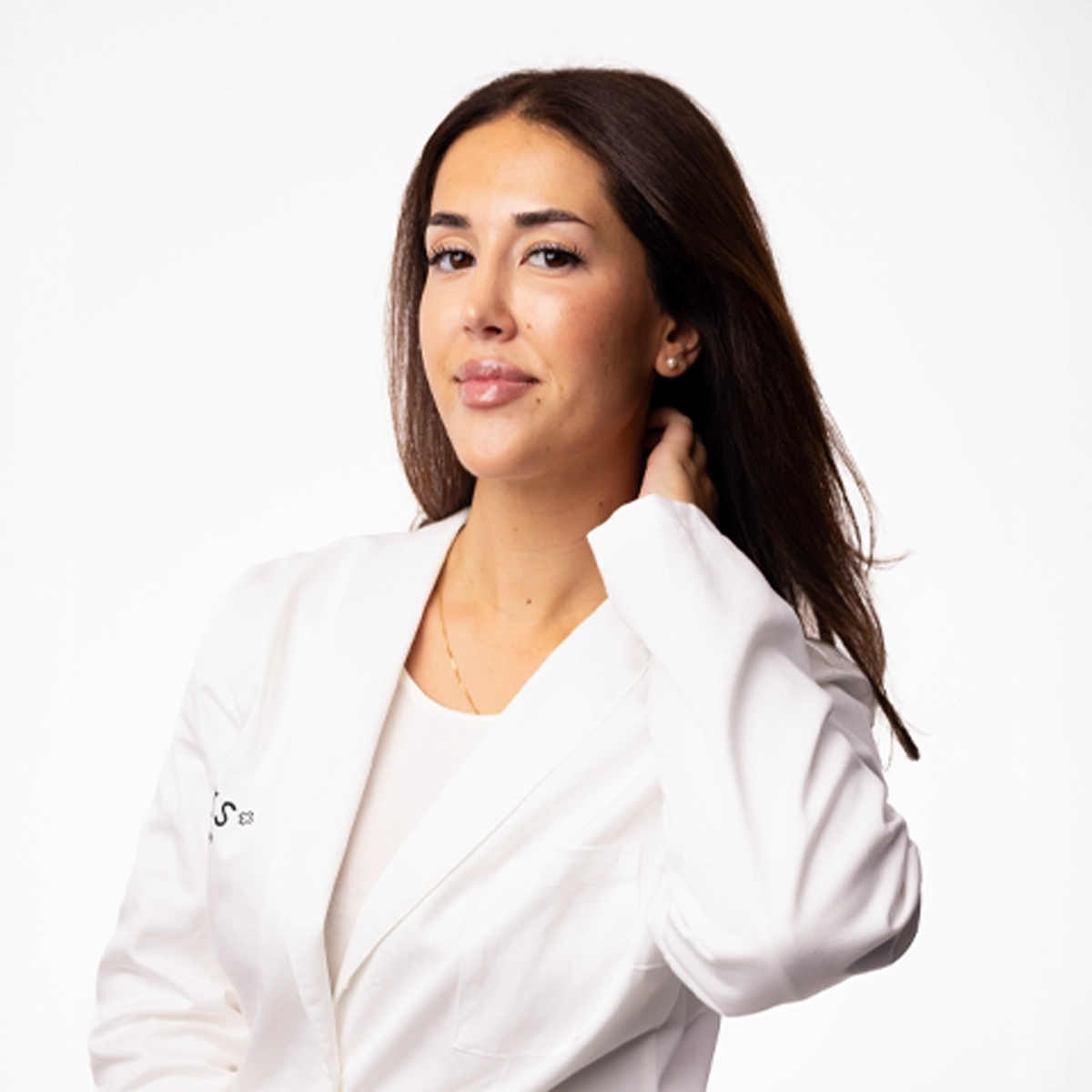 Porträtt av en person med långt, mörkt hår som bär en vit kavaj och en ljus topp. Personen står mot en vit bakgrund och håller ena handen bakom nacken.