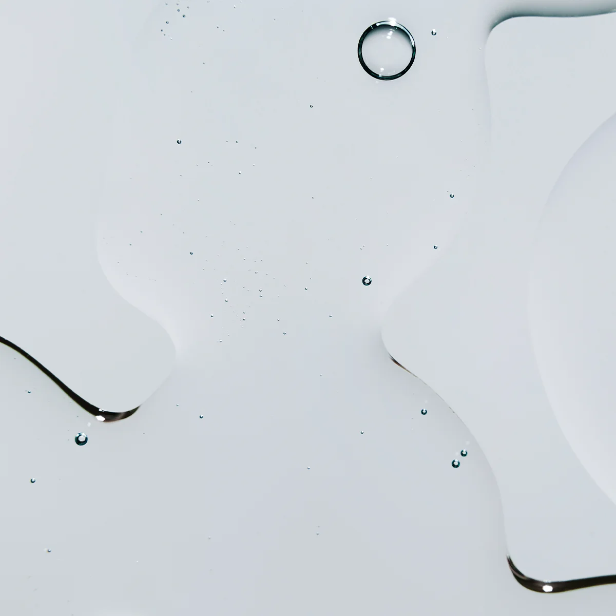 Lähikuva kirkkaasta ihonhoitotekstuurista, joka leviää pehmeisiin muotoihin vaalealla taustalla, pienillä pisaroilla ja ohuilla seerumireunoilla.