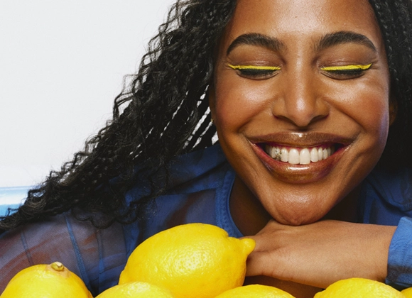 Leende person med flätat hår och färgglad makeup, inklusive gul eyeliner och glansiga bruna läppar. Håller armarna över en hög av citroner.