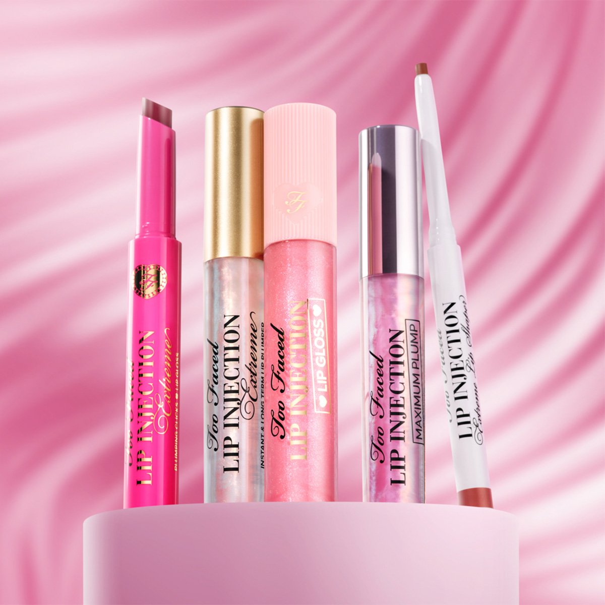 Too Faced -huulituotteita vaaleanpunaisella taustalla, mukana huulikiiltoja ja huulikynä.