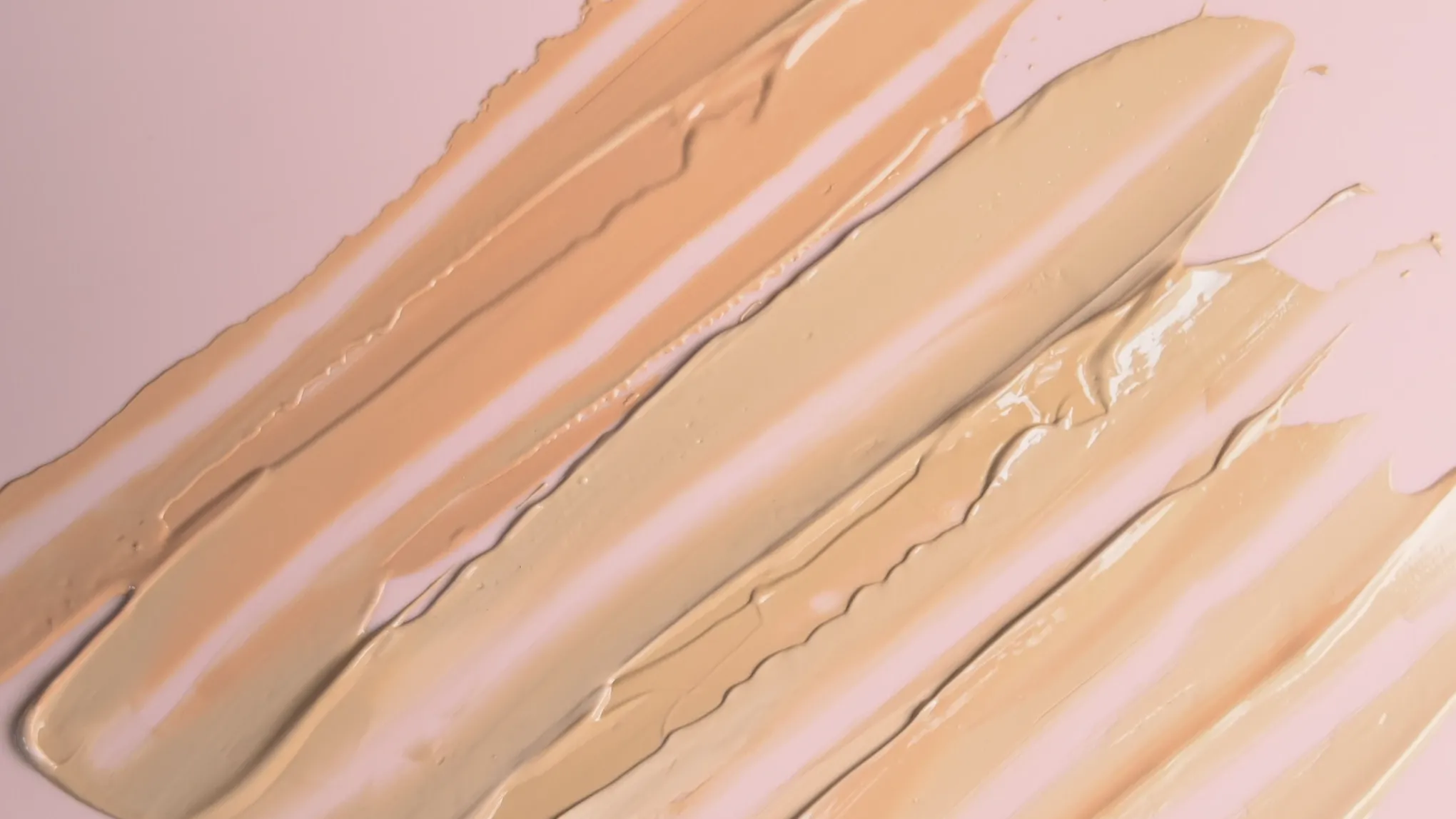 Ulike lyse nyanser av foundation påført i strøk på en lys rosa bakgrunn"