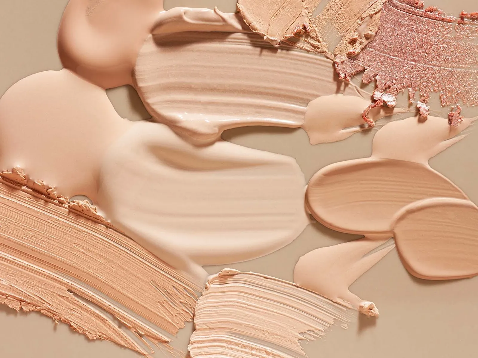 Nærbilde av forskjellige nyanser av foundation og sminke smurt ut på en beige overflate.