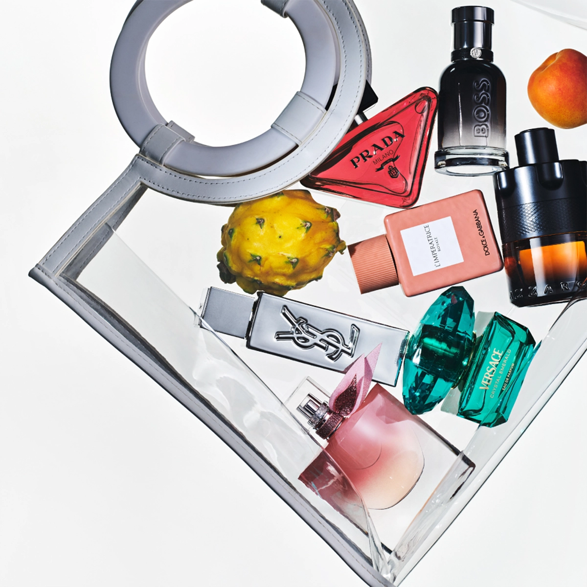 Useita hajuvesipulloja aseteltuna läpinäkyvään käsilaukkuun hedelmien kanssa. Näkyvissä hajuvedet merkeiltä Prada, BOSS, Yves Saint Laurent, Dolce & Gabbana, Versace ja Lancôme eri värisissä lasipulloissa.