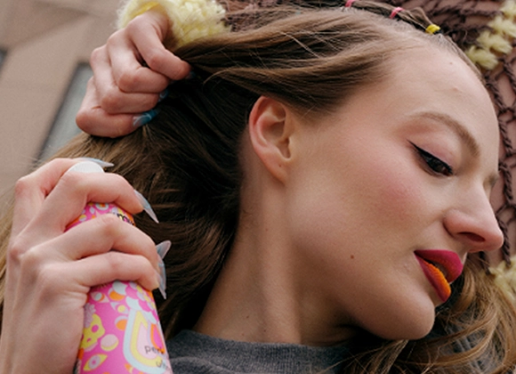 Person med långt hår sprayar torrschampo mot hårbotten. Färgstarkt smink, orange-röda läppar och kreativa naglar syns i en urban miljö.