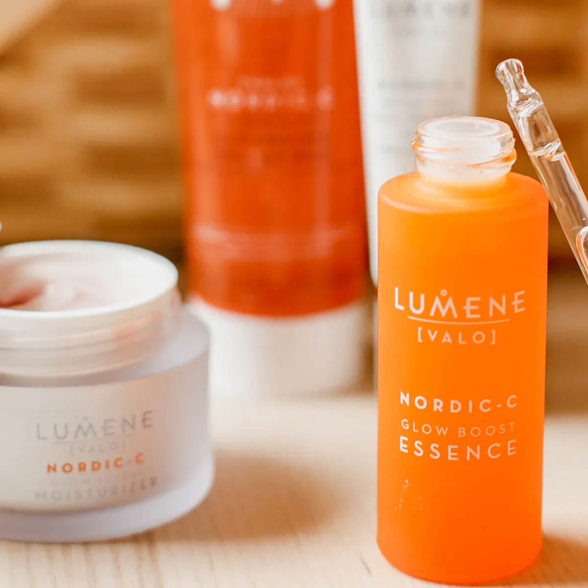 Et nærbilde av Lumene Nordic-C Glow Boost Essence og Nordic-C Glow Reveal Moisturizer. Den oransje flasken med pipette inneholder essensen, mens den hvite krukken inneholder fuktighetskremen. Bakgrunnen viser flere Lumene-produkter.