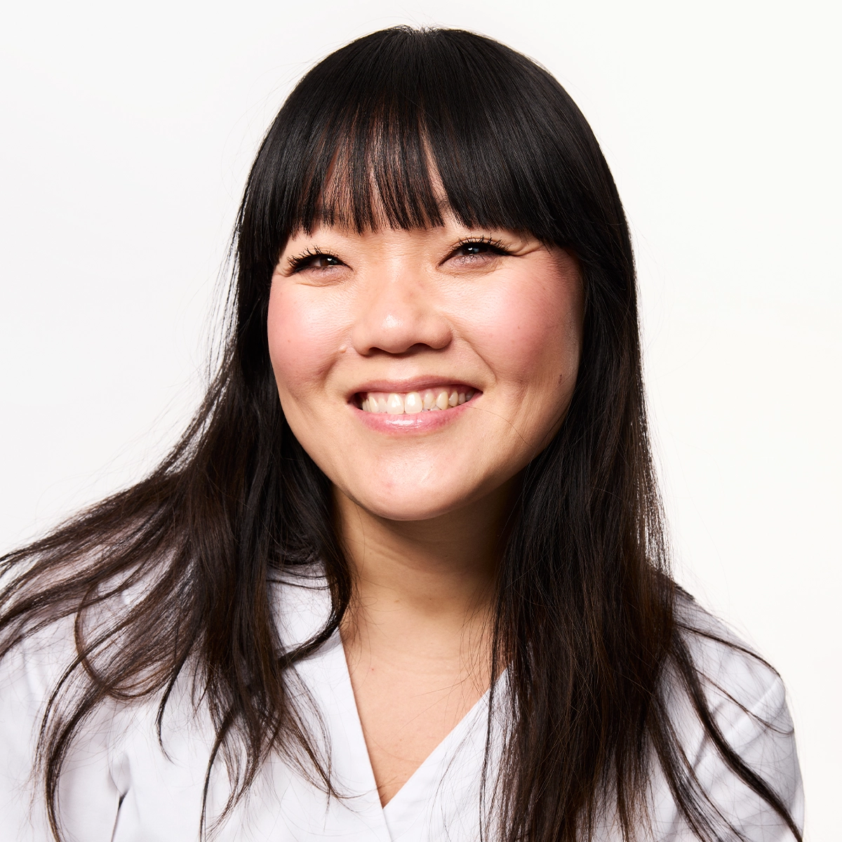 Portrett av en smilende person med langt mørkt hår og rett lugg, fotografert mot lys bakgrunn.