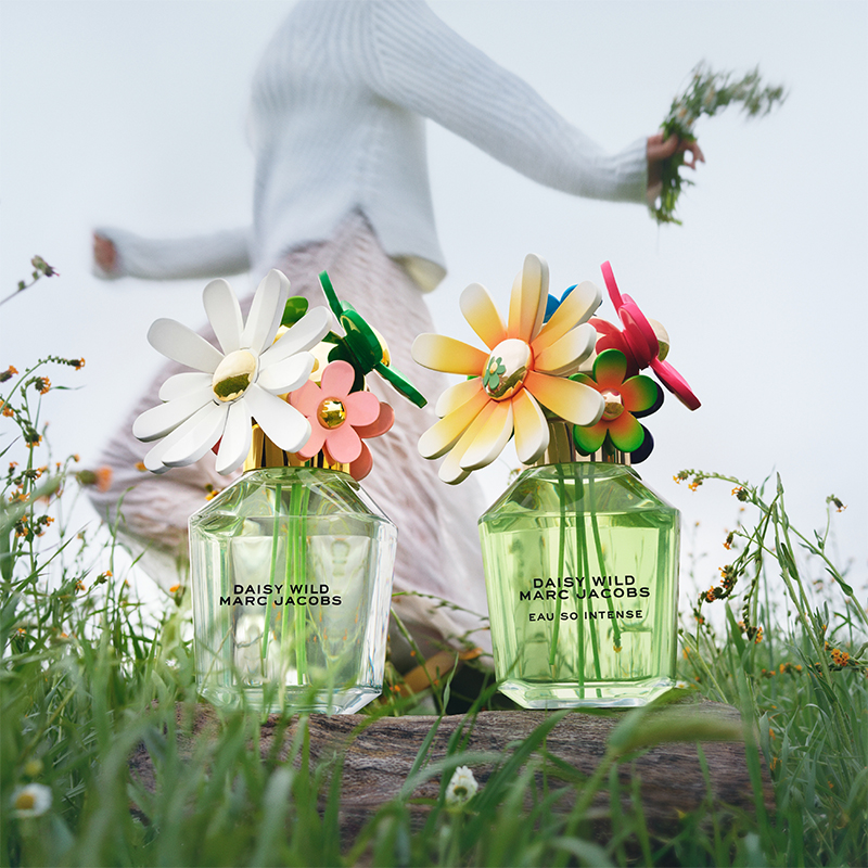 To Daisy Wild Marc Jacobs-parfymeflasker med fargerike blomster som korker. Bak flaskene en person som holder blomster i hånden, kledd i hvit genser og rosa bukser. Bakgrunnen har grønt gress og små blomster.