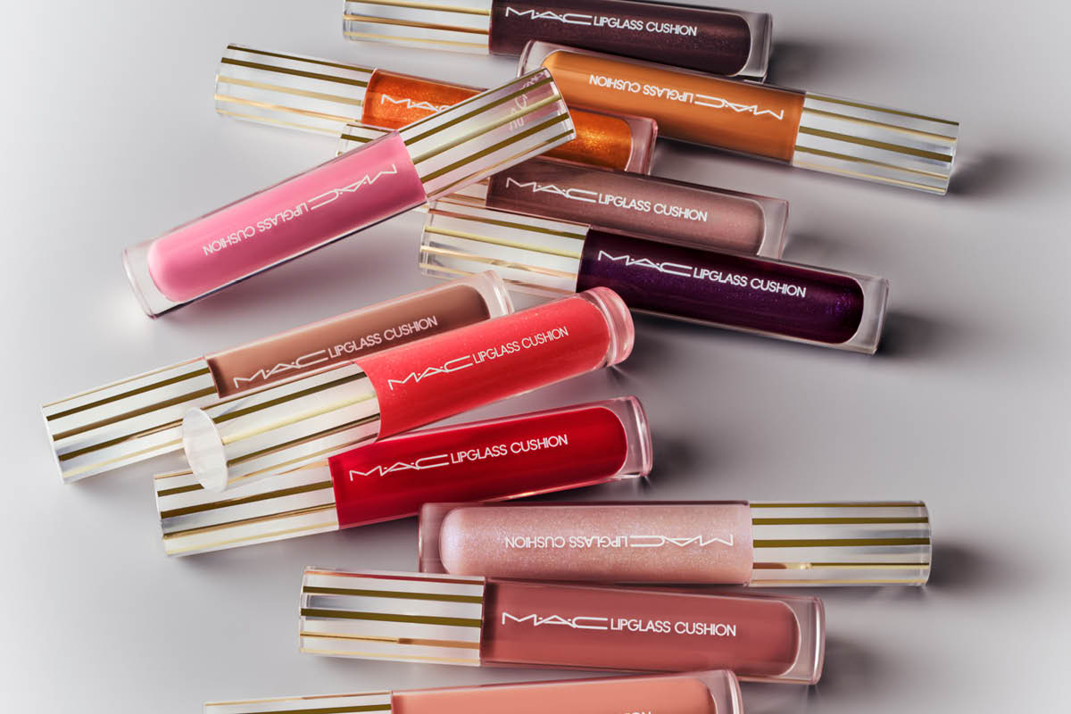 Useita MAC Lipglass Cushion -huulikiiltoja eri sävyissä on aseteltu vaalealle taustalle, kultaraitaisilla korkilla. Tuotteissa lukee “MAC Lipglass Cushion”