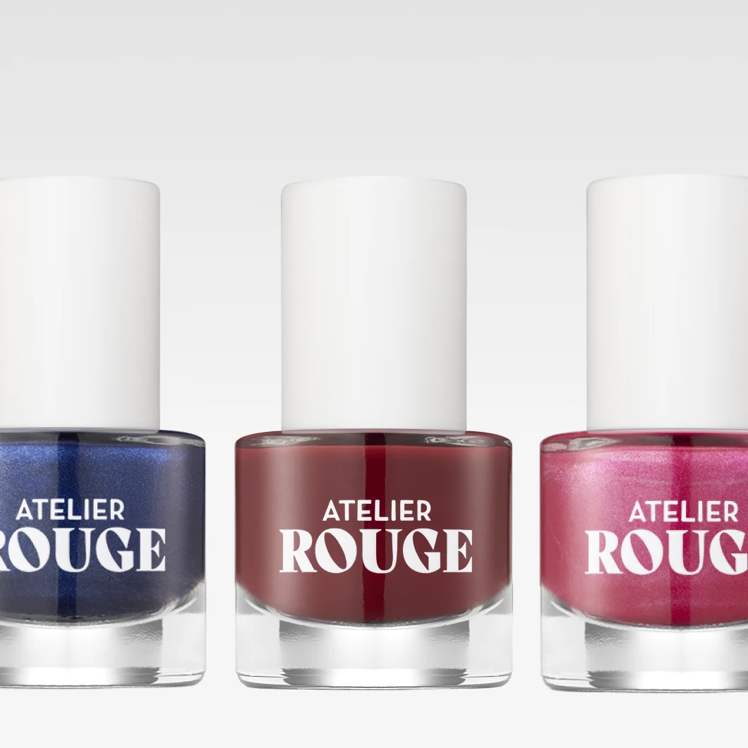 Tre nagellacksflaskor står bredvid varandra mot en ljus bakgrund – i färgerna blå, vinröd och rosa med skimmer. Flaskorna har vita korkar och reflekterar ljuset, vilket skapar ett stilrent, modernt intryck.