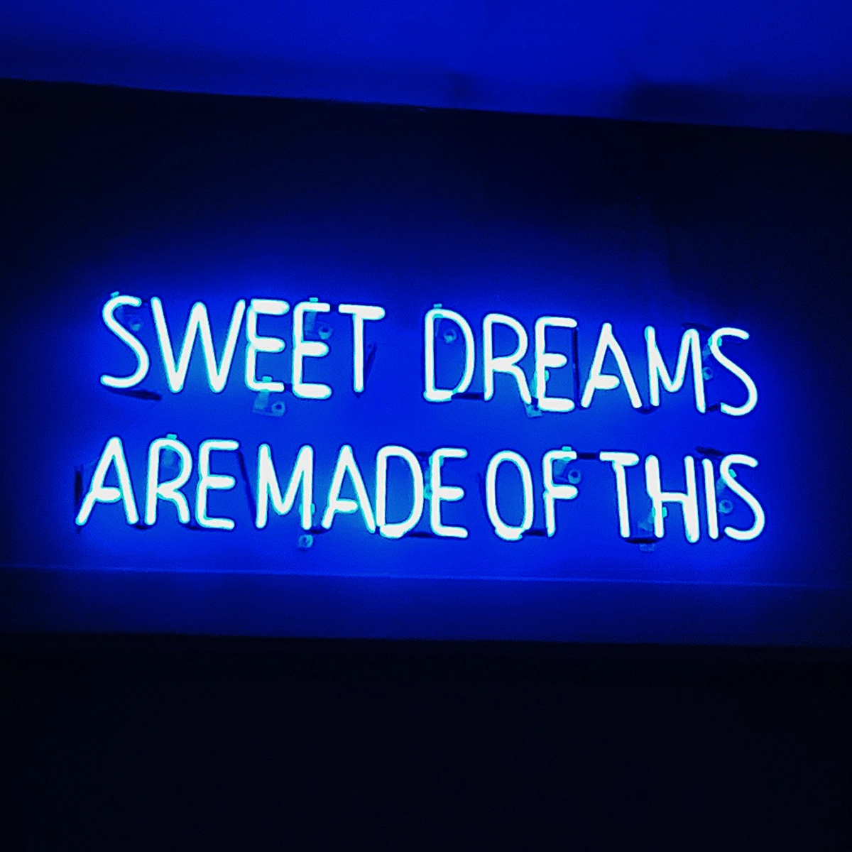 Blått neonskilt på en mørk vegg med den lysende teksten «SWEET DREAMS ARE MADE OF THIS». Skiltet kaster et blått lys på omgivelsene og er bildets sentrale motiv.