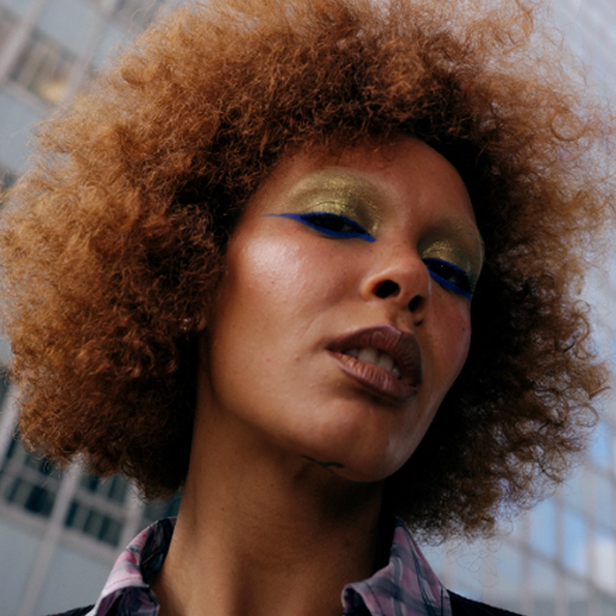Nærbilde av en person med fyldig krøllete hår og makeup med gullfarget øyenskygge og markert blå eyeliner, samt naturlig base og nude lepper.